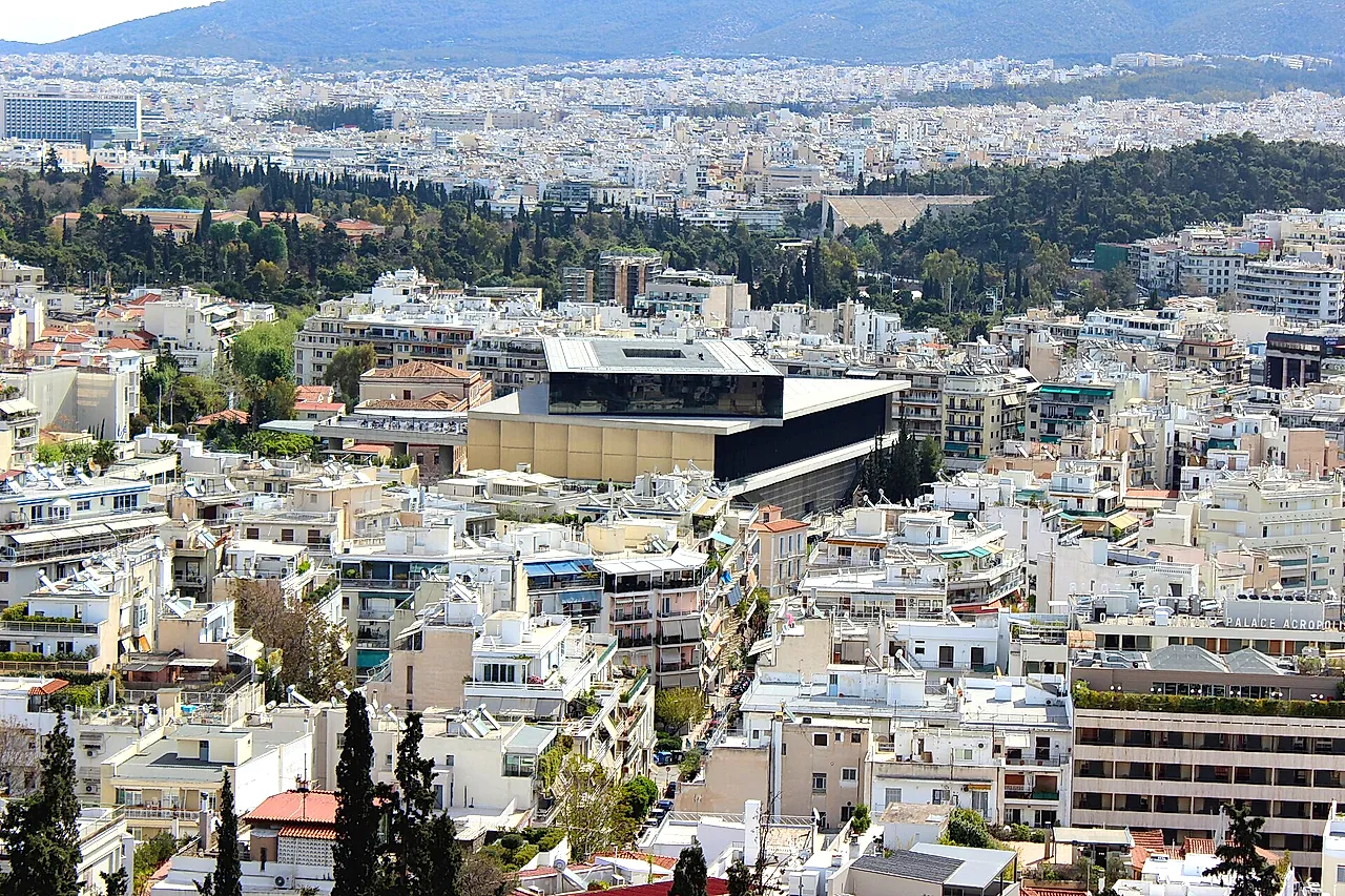 The Acropolis Museum, a modern building of glass and pale stone among the houses of Athens, seen from a hill