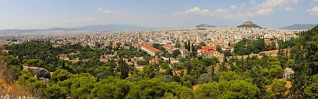 Wide panorama of Athens seen from the Acropolis hill, with pine trees in the foreground and the city stretching to the hills on the horizon